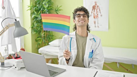 Photo for Young hispanic man doctor using laptop holding rainbow flag at clinic - Royalty Free Image