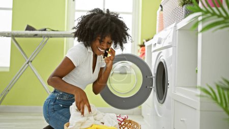 Téléchargez les photos : Femme afro-américaine parlant sur smartphone laver les vêtements souriant à la buanderie - en image libre de droit