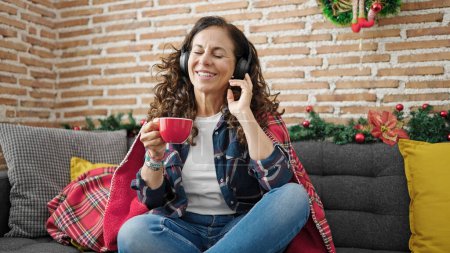 Foto de Mujer hispana de mediana edad escuchando música tomando café celebrando la Navidad en casa - Imagen libre de derechos