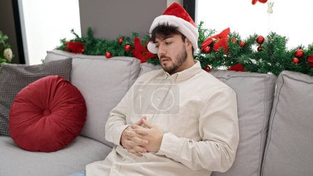 Téléchargez les photos : Jeune homme hispanique souffrant de maux d'estomac portant un chapeau de Noël à la maison - en image libre de droit