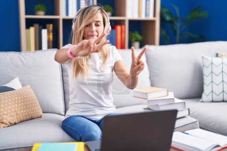 Téléchargez les photos : Jeune femme blonde étudiant à l'aide d'un ordinateur portable à la maison souriant avec la langue montrant les doigts des deux mains faisant signe de victoire. numéro deux. - en image libre de droit