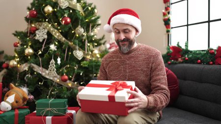Photo for Young bald man wearing christmas hat unpacking gift with surprised face at home - Royalty Free Image