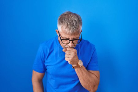 Foto de Hombre hispano con el pelo gris parado sobre fondo azul sintiéndose mal y tosiendo como síntoma de resfriado o bronquitis. concepto de atención sanitaria. - Imagen libre de derechos