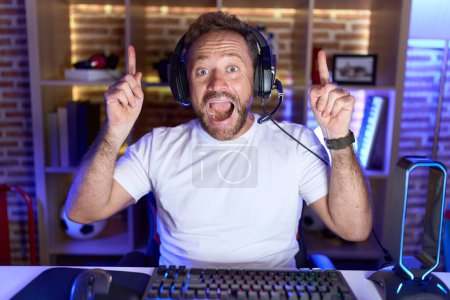 Foto de Hombre de mediana edad con barba jugando videojuegos con auriculares sonriendo sorprendido y sorprendido y apuntando hacia arriba con los dedos y los brazos levantados. - Imagen libre de derechos