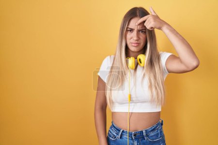 Photo for Young blonde woman standing over yellow background wearing headphones pointing unhappy to pimple on forehead, ugly infection of blackhead. acne and skin problem - Royalty Free Image