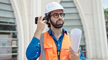 Photo for Young hispanic man architect holding blueprints listening to voice message by smartphone at construction place - Royalty Free Image