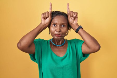 Foto de Mujer africana con rastas de pie sobre fondo amarillo haciendo gesto divertido con el dedo sobre la cabeza como cuernos de toro - Imagen libre de derechos