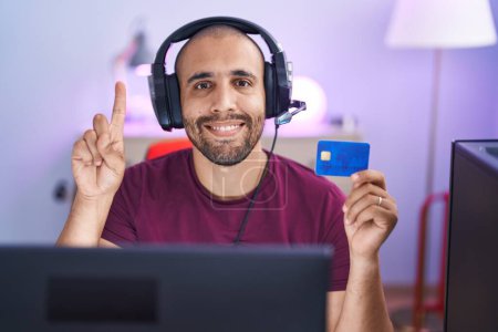 Foto de Hombre hispano con barba haciendo compras en línea con computadora y tarjeta de crédito sonriendo con una idea o pregunta apuntando con el dedo con la cara feliz, número uno - Imagen libre de derechos