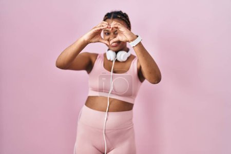 Téléchargez les photos : Femme afro-américaine avec des tresses portant des vêtements de sport et des écouteurs faisant forme de coeur avec la main et les doigts souriant en regardant à travers signe - en image libre de droit