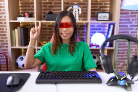 Foto de Mujer china de mediana edad que usa gafas de realidad virtual que muestran y señalan con el dedo número uno mientras sonríe confiada y feliz. - Imagen libre de derechos