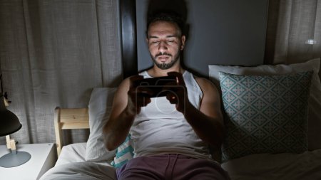 Photo for Young arab man playing video game sitting on bed at bedroom - Royalty Free Image