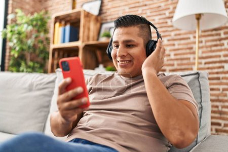 Téléchargez les photos : Jeune homme latin écoutant de la musique assis sur le canapé à la maison - en image libre de droit