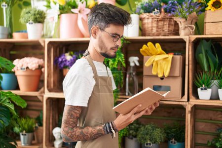 Téléchargez les photos : Jeune homme hispanique fleuriste lecture carnet avec expression détendue à la boutique de fleurs - en image libre de droit