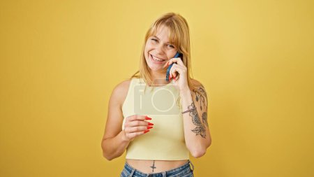 Téléchargez les photos : Jeune femme blonde souriante confiante parlant sur le smartphone sur fond jaune isolé - en image libre de droit