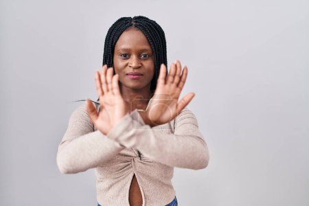 Foto de Mujer africana con trenzas de pie sobre fondo blanco expresión rechazo cruzando brazos y palmas haciendo signo negativo, cara enojada - Imagen libre de derechos