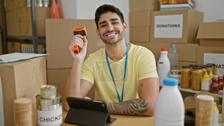 Foto de Joven hispano voluntario sonriendo confiado sosteniendo lector de códigos de barras en el centro de caridad - Imagen libre de derechos