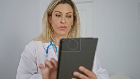 Foto de Mujer joven rubia médico usando touchpad ith cara seria en la sala de espera clínica - Imagen libre de derechos