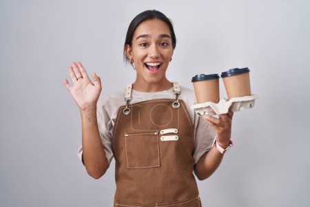 Foto de Mujer hispana joven vistiendo delantal de camarera profesional sosteniendo café renunciando a decir hola feliz y sonriente, gesto de bienvenida amistoso - Imagen libre de derechos