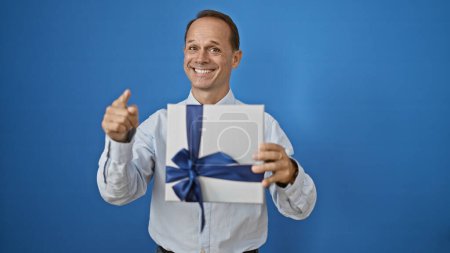 Foto de Alegre hombre de mediana edad, rebosante confianza, sosteniendo una sorpresa de cumpleaños, sonriendo alegremente y señalando a la cámara contra un fondo de pared blanco aislado - Imagen libre de derechos