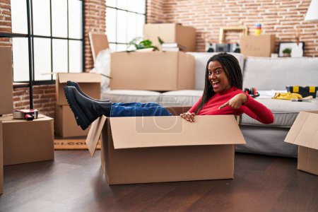 Téléchargez les photos : Jeune afro-américain avec des tresses se déplaçant vers une nouvelle maison à l'intérieur d'une boîte en carton souriant heureux pointant avec la main et le doigt - en image libre de droit