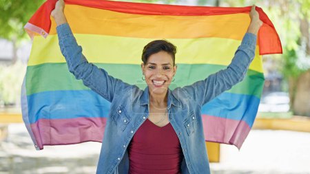 Foto de Joven hermosa mujer hispana sonriendo confiada sosteniendo la bandera del arco iris en el parque - Imagen libre de derechos