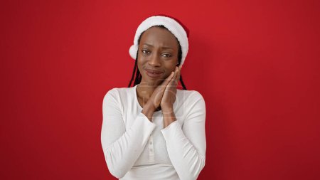 Téléchargez les photos : Femme afro-américaine souriante et confiante portant un chapeau de Noël sur fond rouge isolé - en image libre de droit