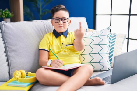 Photo for Young hispanic kid doing homework sitting on the sofa showing middle finger, impolite and rude fuck off expression - Royalty Free Image