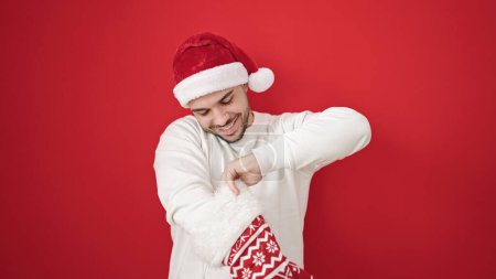 Téléchargez les photos : Jeune homme hispanique portant un chapeau de Noël regardant à l'intérieur de la chaussette sur fond rouge isolé - en image libre de droit