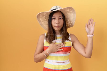 Téléchargez les photos : Femme chinoise du Moyen Âge portant un chapeau d'été sur fond jaune jurant avec la main sur la poitrine et la paume ouverte, faisant un serment de fidélité - en image libre de droit