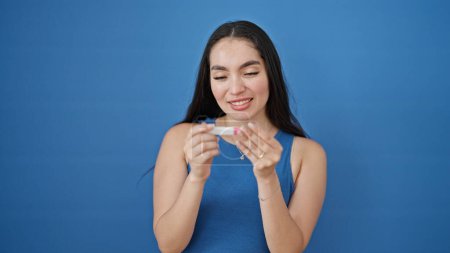 Foto de Joven mujer hispana hermosa sosteniendo prueba de embarazo sonriendo sobre fondo azul aislado - Imagen libre de derechos