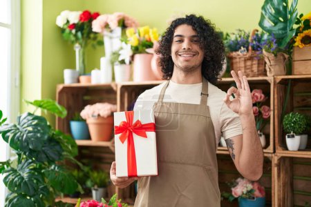 Photo for Hispanic man with curly hair working at florist shop holding gift doing ok sign with fingers, smiling friendly gesturing excellent symbol - Royalty Free Image