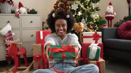 Photo for African american woman wearing reindeer ears holding christmas gift at home - Royalty Free Image