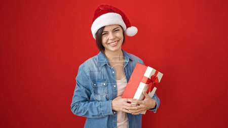 Téléchargez les photos : Jeune femme caucasienne souriante confiante portant un chapeau de Noël sur fond rouge isolé - en image libre de droit