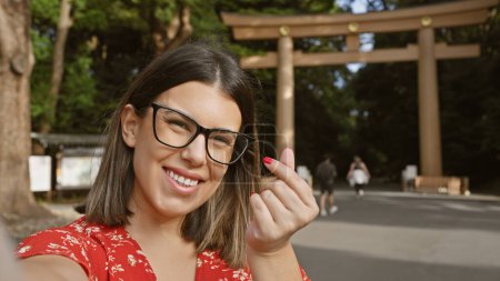 Photo for Cheerful young hispanic woman in glasses captures her heart-shaped hand sign in a selfie at japan's iconic meiji temple, exuding love and happiness in this beautiful picture - Royalty Free Image