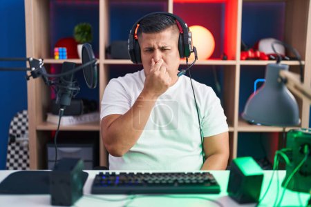 Foto de Young hispanic man playing video games smelling something stinky and disgusting, intolerable smell, holding breath with fingers on nose. bad smell - Imagen libre de derechos