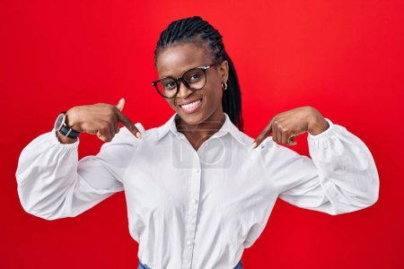 Téléchargez les photos : Femme africaine avec des tresses debout sur fond rouge regardant confiant avec le sourire sur le visage, se montrant du doigt fier et heureux. - en image libre de droit