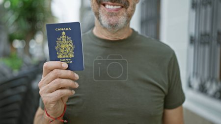 Téléchargez les photos : Homme confiant et souriant montrant son passeport canadien dans la rue de la ville, se prélassant de la lumière du soleil, du bonheur rayonnant de ses cheveux gris et de sa barbe mature - en image libre de droit