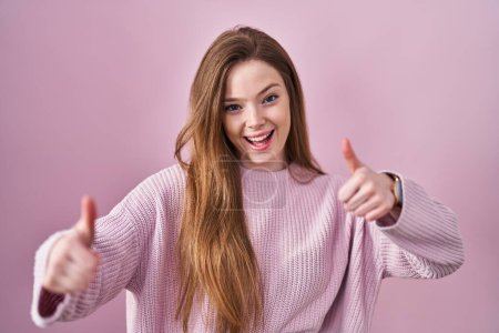 Téléchargez les photos : Jeune femme caucasienne debout sur fond rose approuvant faire un geste positif avec la main, pouces levés souriant et heureux pour le succès. geste gagnant. - en image libre de droit