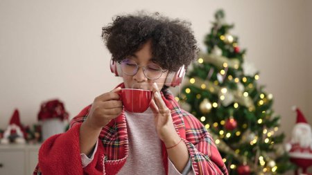 Photo for Young african american woman drinking coffee standing by christmas tree dancing at home - Royalty Free Image