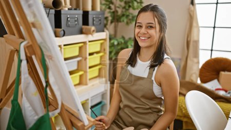 Foto de Sonriente joven artista hispana, una mujer hermosa y confiada dibujando en su estudio de arte, disfrutando del hobby creativo - Imagen libre de derechos