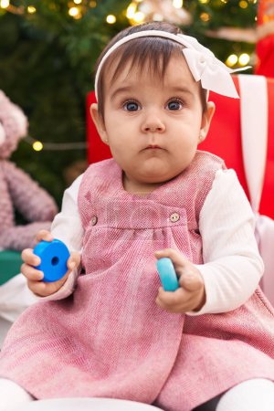 Téléchargez les photos : Adorable bébé hispanique tenant cerceaux jouet assis sur le sol près de l'arbre de Noël à la maison - en image libre de droit