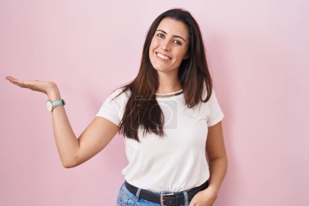 Téléchargez les photos : Jeune femme brune debout sur fond rose souriant gai présenter et pointant avec la paume de la main en regardant la caméra. - en image libre de droit