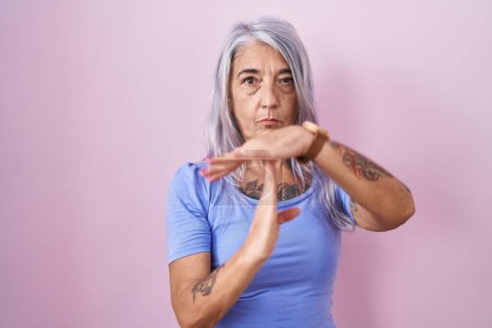 Foto de Mujer de mediana edad con tatuajes de pie sobre fondo rosa haciendo tiempo fuera gesto con las manos, rostro frustrado y grave - Imagen libre de derechos