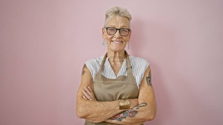 Photo for Confident senior grey-haired woman waitress, smiling and standing firm with arms crossed over isolated pink background - Royalty Free Image