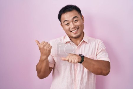Foto de Chino joven de pie sobre fondo rosa apuntando hacia atrás con la mano y los pulgares hacia arriba, sonriendo confiado - Imagen libre de derechos