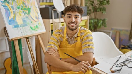 Téléchargez les photos : Confiant, souriant jeune artiste arabe assis avec les bras croisés dans son atelier d'art, embrassant joyeusement son passe-temps de peinture - en image libre de droit