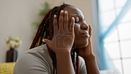 Foto de Deprimida mujer afroamericana con trenzas sentada sola en el sofá en casa, expresión infeliz revela dolor de cabeza y estrés - Imagen libre de derechos