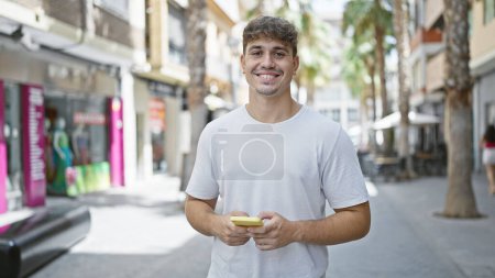 Téléchargez les photos : Jeune homme hispanique utilisant smartphone souriant à la rue - en image libre de droit