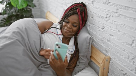 Foto de Relajada mujer afroamericana acostada en la cama, enviando mensajes de texto cómodamente en el teléfono inteligente en su acogedor dormitorio - Imagen libre de derechos