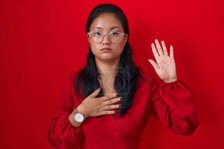 Téléchargez les photos : Asiatique jeune femme debout sur fond rouge jurant avec la main sur la poitrine et la paume ouverte, faisant un serment de fidélité promesse - en image libre de droit